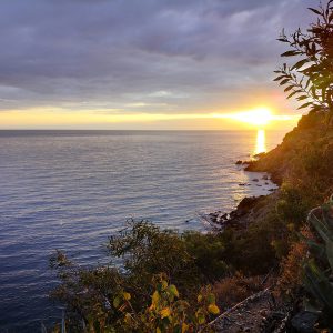 Naplemente a tengerparton. Jobb oldalt a dombok és sziklák látszanaka, a fotó nagyobb részében pedig a tengert láthatjuk a naplemente fényében.